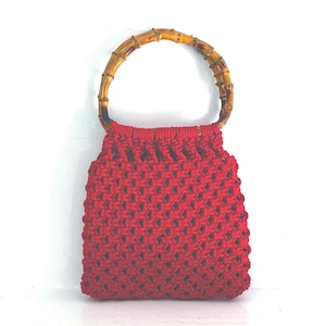 May include: A bright red handbag crafted with a knotted macrame cord pattern, featuring a rounded handle made from natural bamboo segments. The bag has a square-shaped body with a dense, textured weave and a solid, vibrant red color throughout.