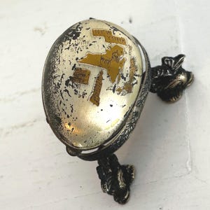 May include: Vintage silver-toned compact mirror with a New York state map design. The map features gold-colored accents and iconic landmarks. The compact has a decorative floral design on the handle.