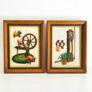 May include: Two framed embroidery pieces with a wooden frame. The left piece features a spinning wheel with a basket of apples. The right piece features a grandfather clock with a pendulum.