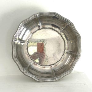 May include: A round, polished silver-toned metal bowl with a scalloped, wavy edge and a smooth, reflective interior surface. The bowl features a multi-tiered, stepped rim design that creates a series of concentric ridges around the perimeter.