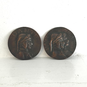 May include: Two bronze-colored round medallions. Each features a profile of a person wearing a hat, with the word "DANTE" around the edge. One medallion shows a single profile, the other shows two profiles. The medallions are on a white surface.