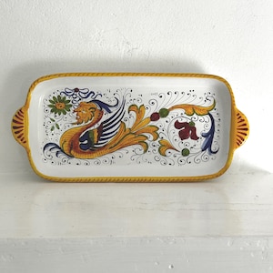 May include: A rectangular ceramic serving tray with a white base and a yellow rim. The center features a hand-painted design of a mythical creature with yellow, blue, and green accents, surrounded by intricate black scrollwork and floral motifs.