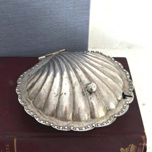 May include: A silver-toned metal clam shell dish with a hinged lid and a small spherical knob. The exterior features a ribbed, scalloped design with a decorative beaded border along the edge. The metal surface shows signs of wear and patina.
