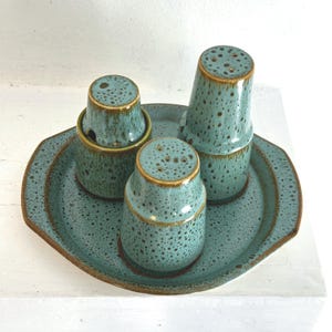 May include: A teal ceramic salt and pepper set with a matching tray. The set includes two shakers, a lidded container, and a tray. The pieces have a speckled pattern and a brown rim. The tray is approximately 8 inches in diameter.