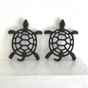 May include: Two black metal turtle figurines with openwork shell designs. The turtles have outstretched flippers and a small head. They are displayed on a white surface against a white wall.