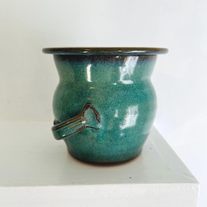 May include: A teal ceramic planter with a unique handle. The planter has a glazed finish and a slightly rounded shape.