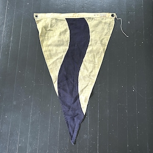 May include: A triangular pennant flag made of beige fabric with a central vertical wavy dark blue stripe. The top edge features a reinforced header with two metal grommets and a small white tag with printed text, including the word "Standard".