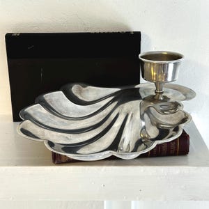 May include: A silver-toned metal serving dish shaped like a large scallop shell with a smooth, reflective surface. Attached to the right side of the shell is a small, cylindrical cup on a pedestal base, designed for holding a dip or small condiment.