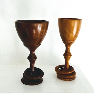 May include: Two wooden goblets with a dark brown and light brown finish. The goblets have a unique stem design with a series of rings at the base. The cups are made of wood and have a smooth, polished appearance.