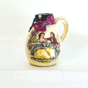 May include: A cream-colored ceramic pitcher with a handle, featuring a painted scene of two figures in period clothing. The pitcher is adorned with a black lid, a purple bow, and blue ribbon. The overall design is vintage and decorative.
