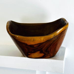 May include: A handcrafted wooden bowl with a natural, organic shape. The bowl features rich brown and amber tones, showcasing the wood grain. The interior is a darker shade of brown, creating a contrast with the lighter exterior. The bowl is displayed on a white shelf.
