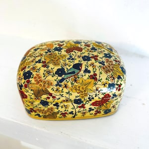 May include: A decorative, hand-painted box with a rounded shape. The box features a yellow background with a floral and bird design in shades of blue, red, yellow, and gold. The box is sitting on a white surface.