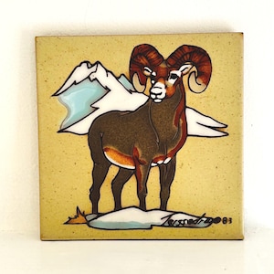 May include: A ceramic tile with a hand-painted design of a brown bighorn sheep with large curled horns standing on a snowy mountain scene. The tile has a yellow background and the artist's signature and the year '83' are visible in the bottom right corner.