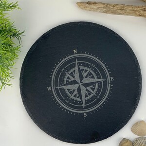 Könnte beinhalten: Runder Untersetzer aus schwarzem Schiefer mit einem gravierten Kompass-Design in Silber. Der Kompass zeigt Norden, Süden, Osten und Westen.
