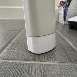Op de afbeelding: Een deel van een lichtgekleurd, rechthoekig object met een witte, getextureerde basis. Het object staat op een grijze tegelvloer met houtlook. De basis heeft een afgeronde, gebogen vorm.