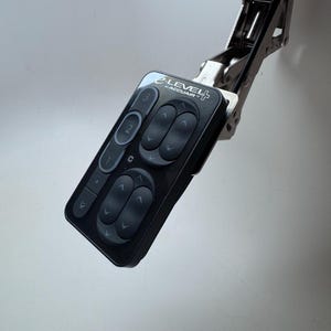 Può includere: Un pannello di controllo nero con la scritta "e-LEVEL by ACCUAIR" e diversi pulsanti. Il pannello è fissato a una staffa metallica argentata. I pulsanti sono etichettati con numeri e frecce.