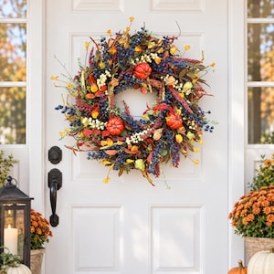 Puede incluir: Una corona de otoño circular que presenta pequeñas calabazas naranjas, racimos de bayas azul oscuro, bayas color crema y varias hojas secas en tonos amarillos, rojos y marrones, dispuestas sobre un marco circular para colgar en una puerta.