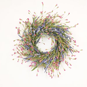 Puede incluir: Una corona circular compuesta por elementos florales de lavanda, rosa y verde. La corona presenta una mezcla de flores de color lavanda, pequeños capullos rosas y follaje verde, creando una apariencia natural y vibrante. Ideal para la decoración del hogar.