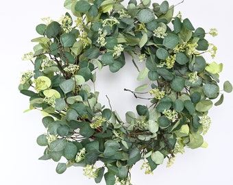 Könnte beinhalten: Ein grüner Eukalyptus-Kranz mit kleinen weißen Blüten. Der Kranz ist aus künstlichem Grün hergestellt und perfekt, um Ihrem Zuhause einen Hauch von Natur zu verleihen.