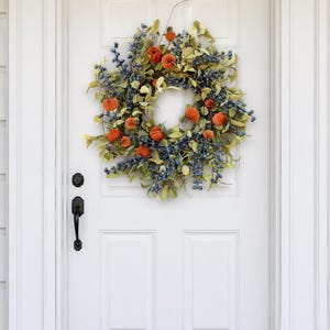 Peut inclure: Une couronne circulaire composée de petites citrouilles orange entremêlées de grappes de baies bleues et de feuilles vert clair. La couronne est fixée sur une porte blanche à panneaux dotée d'une poignée et d'un verrou noirs.