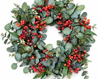 May include: A circular wreath featuring a dense arrangement of muted green eucalyptus leaves and clusters of bright red berries. The foliage includes rounded and elongated leaf shapes interspersed with small, light-coloured floral buds throughout.