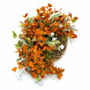 Puede incluir: Una corona decorativa que presenta un arreglo denso de hojas otoñales de color naranja vibrante y follaje verde, acentuado con pequeñas flores de color azul claro. El conjunto botánico está sujeto a una base circular hecha de ramas marrones entrelazadas.