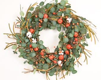 May include: A circular wreath featuring green eucalyptus leaves, thin brown twigs, small orange pumpkins, and white cotton bolls. The arrangement is dense and textured, combining natural elements in a round decorative display for wall or door hanging.