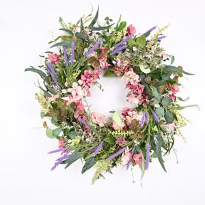 May include: A circular floral wreath featuring a variety of faux flowers and greenery. The wreath includes pink and purple flowers, green leaves, and sprigs of lavender. The wreath is set against a white background.