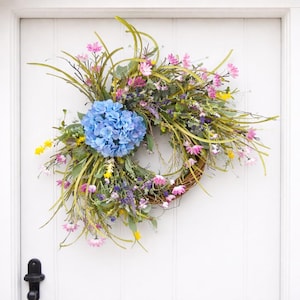 May include: A vibrant floral wreath with a woven base, adorned with a large blue hydrangea, pink daisies, yellow accents, and various greenery. The wreath is displayed on a white door, creating a welcoming spring or summer aesthetic.