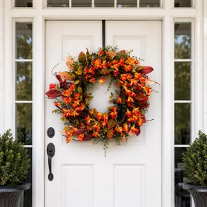 May include: A circular wreath featuring a dense arrangement of artificial orange and yellow flowers, dark red and green leaves, and small dark berries. The wreath is mounted on a white door with a black metal hanger and a black door handle.
