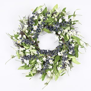 Puede incluir: Una corona hecha de hojas verdes y flores blancas con bayas azules. La corona es perfecta para la decoración de primavera o verano.