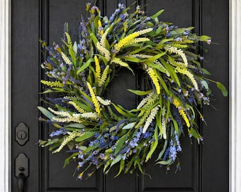May include: A vibrant floral wreath with a mix of yellow, white, and purple flowers and green leaves. The wreath is displayed on a black door, creating a striking contrast. A decorative accent for any home.