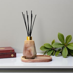 May include: A decorative reed diffuser set. It features a geometric, marbled-effect bottle with a gold-colored collar, black reeds, and a matching oval tray. The color scheme includes brown and beige tones. The diffuser is designed for home fragrance.