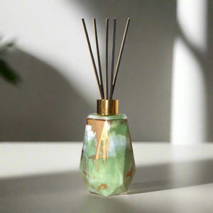 May include: A green glass diffuser bottle with gold accents and black diffuser sticks. The bottle has a geometric shape and a white and green marbled design.