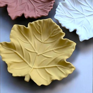 May include: Three ceramic leaf-shaped serving dishes in different colours: yellow, pink and white. The dishes are made to look like leaves with detailed veins and edges.