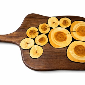 May include: A dark brown wooden serving board with a handle. The board features inlaid circular wood slices with light brown and yellow tones. The wood slices are arranged on the board's surface, creating a decorative design.