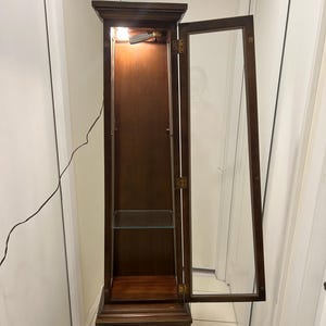 May include: A tall, dark wood display cabinet with a glass door, open to the right. The cabinet has a light inside, a glass shelf, and a decorative base with a drawer and a round handle. The cabinet is in a room with white walls.