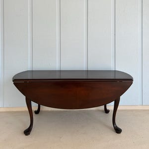 May include: A dark brown, oval-shaped drop-leaf table with curved legs. The table is made of wood and has a polished finish. A framed floral painting hangs on the light blue wall behind the table.