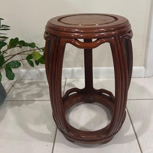 May include: A dark brown, wooden plant stand with a round top and a decorative, openwork design. The stand is approximately 18 inches tall and features intricate, curved supports and a circular base. The wood has a rich, polished finish.