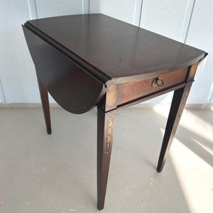 May include: A dark brown, antique drop-leaf table with a drawer and decorative inlay. The table has a rounded top when the leaf is down and stands on four slender legs. The table is approximately 30 inches long when the leaf is up.