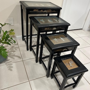 May include: Set of four nesting tables with black frames and glass tops. Each table features a decorative panel under the glass, with a different design. The tables are arranged in a descending size order.