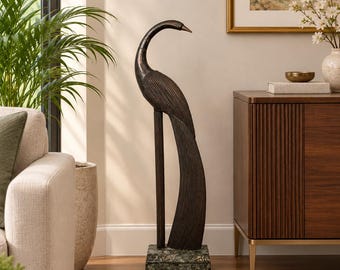 May include: A tall, dark bronze-toned sculpture of a standing bird with a long, curved neck and textured, linear plumage details. The bird figure is mounted on a rectangular, mottled green stone base, creating a vertical, slender silhouette.