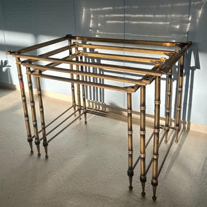 May include: A vintage brass side table with a bamboo-style frame. The table has a rectangular top and a lower shelf, supported by four legs. The legs are adorned with dark accents and end in small, rounded feet.