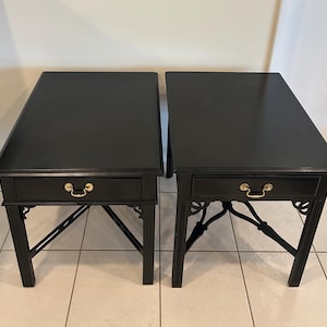 May include: Two black side tables with a drawer and gold-colored hardware. The tables have a rectangular top and decorative legs. The drawer has a gold-colored handle.