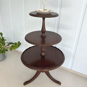 May include: A three-tiered, dark brown wooden serving stand with a decorative scalloped edge on the bottom tier. The top tier holds a small, round plate with a gold handle. The stand is approximately 30 inches tall.