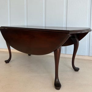 May include: A dark brown, antique drop-leaf table with a semi-circular top and curved legs. The table is made of wood and has a polished finish. The table's dimensions are approximately 36 inches long, 24 inches wide, and 28 inches high.