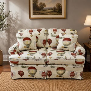 May include: A two-seater loveseat upholstered in an off-white fabric featuring a repeating pattern of vintage-style hot air balloons in shades of red, brown, and tan. The sofa includes two matching square throw pillows and rolled armrests.