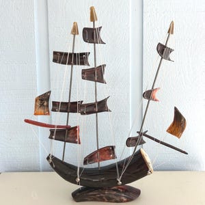 May include: A decorative wooden ship model with dark brown hull and sails. The sails are a mix of brown and orange, and the masts are topped with gold accents. The ship is displayed on a light-colored surface against a light blue background.