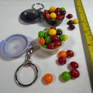 May include: Two small, round, clear plastic containers attached to metal keychains, one with a light purple lid and one with a dark gray lid. Both are filled with colorful, round, fruit-flavored candies. A yellow measuring tape shows 9 inches.
