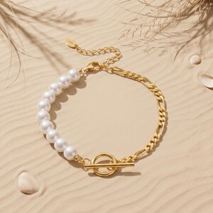 May include: A gold-toned bracelet featuring a half-strand of white spherical pearls and a half-strand of gold-toned metal figaro chain. The bracelet includes a gold-toned toggle clasp closure with a circular ring and a horizontal bar component.